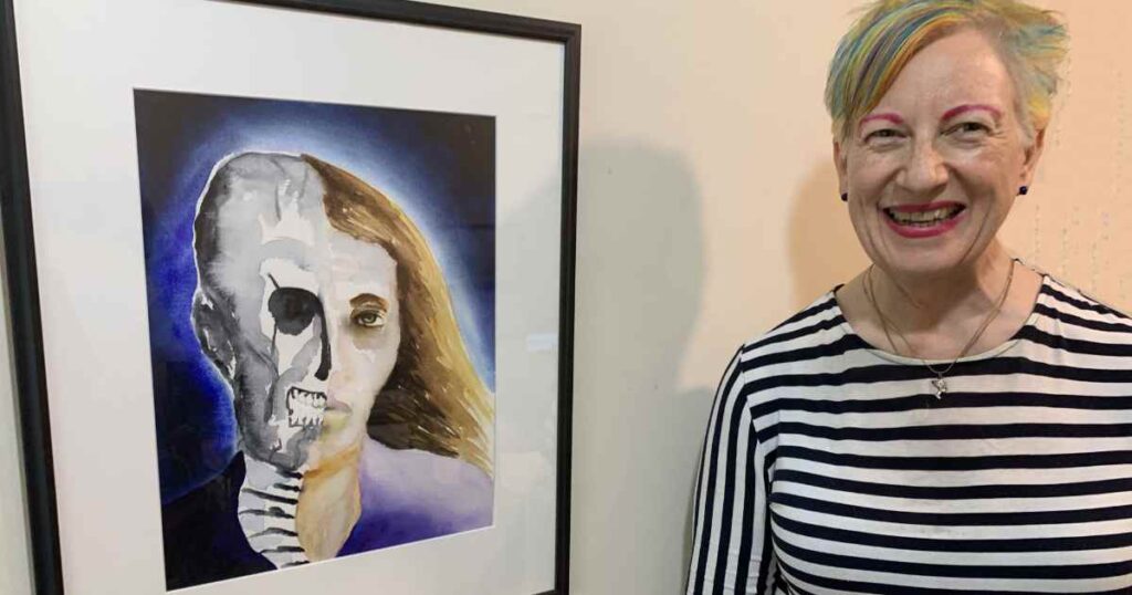 Artist Anne Nordhaus-Bike next to her framed watercolor portrait with half skull, half woman with long hair